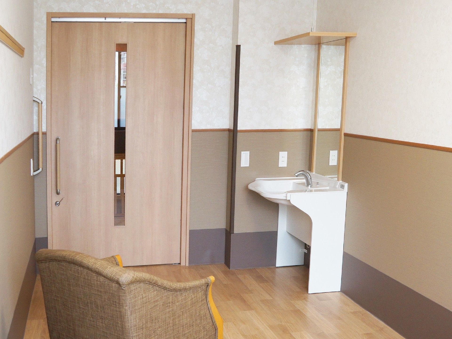 居室の窓側からのショット。出入り口付近に洗面台があり、居室中央には一人用のいすが置かれている。