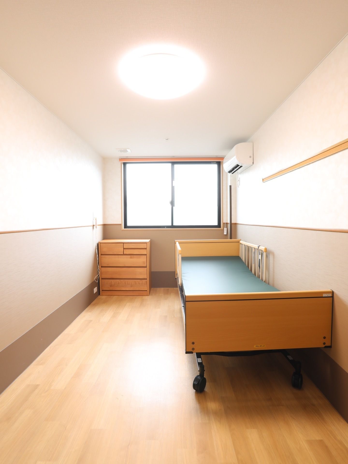 居室の突き当りに窓があり、外光が入っている。介護用ベッドが一台と木製のチェストが置かれ、壁にはエアコンがある。