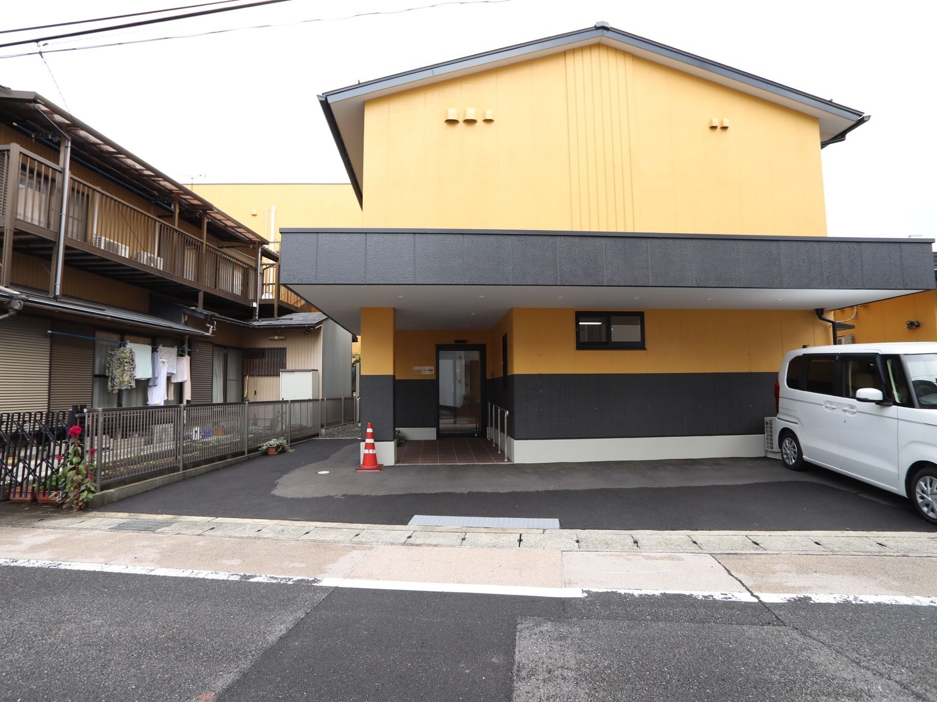 アスファルトの敷地に、2階建ての住宅が建っている。道路と敷地の間は段差がなく、玄関アプローチは屋根が付いている。