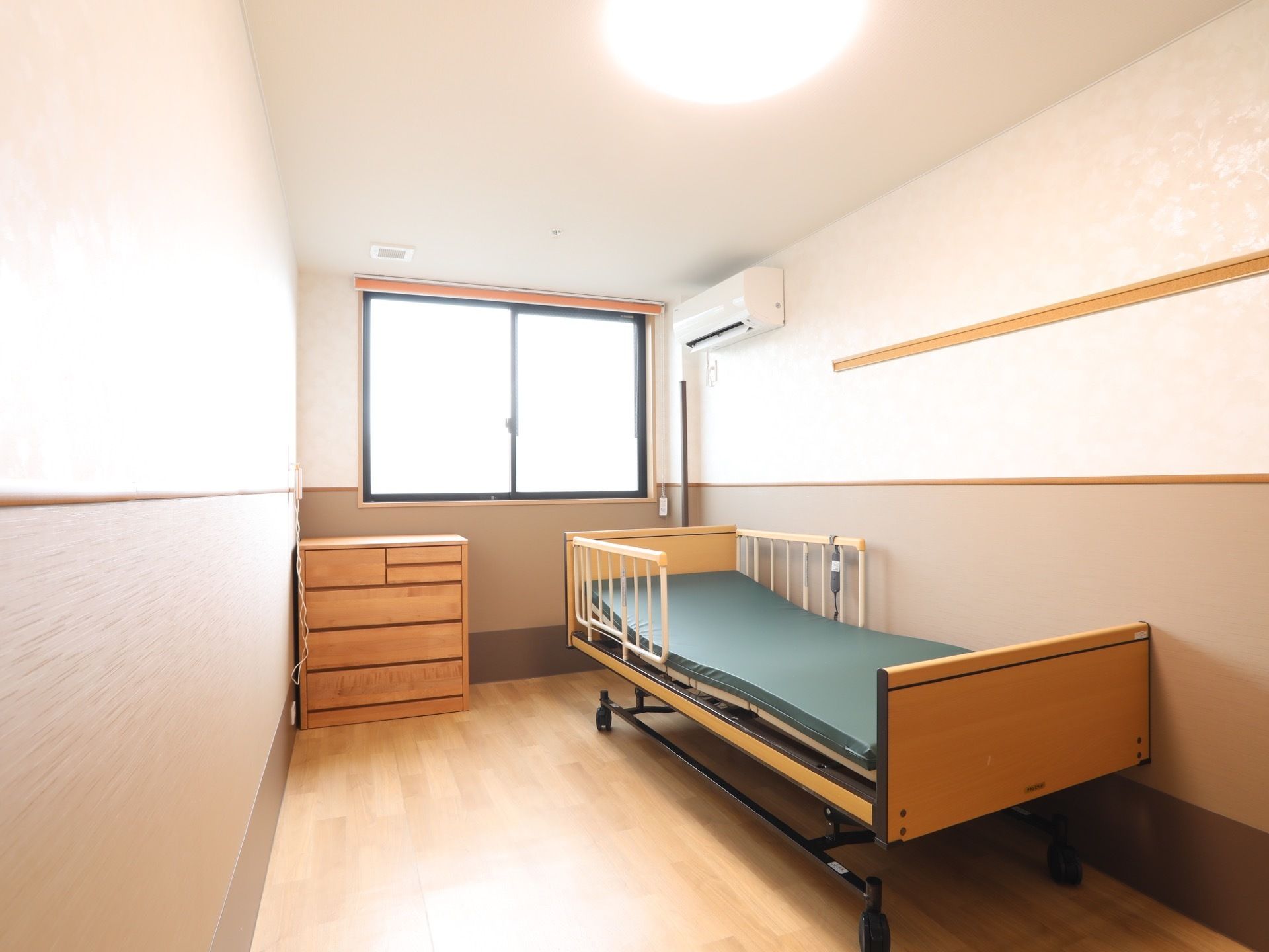 窓の付いたフローリングの居室に、キャスター付きの電動ベッドが一台置かれている。ベッドの横には背の低いチェストもある。