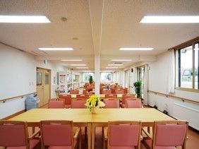 白い壁に木製のダイニングテーブル、テーブルにはピンクのいすが設置されている。観葉植物や花が飾られ家庭的な雰囲気。