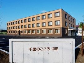 ３階建ての建物は１階部分がダークブラウン、２階より上がライトブラウン。広い駐車場もある。