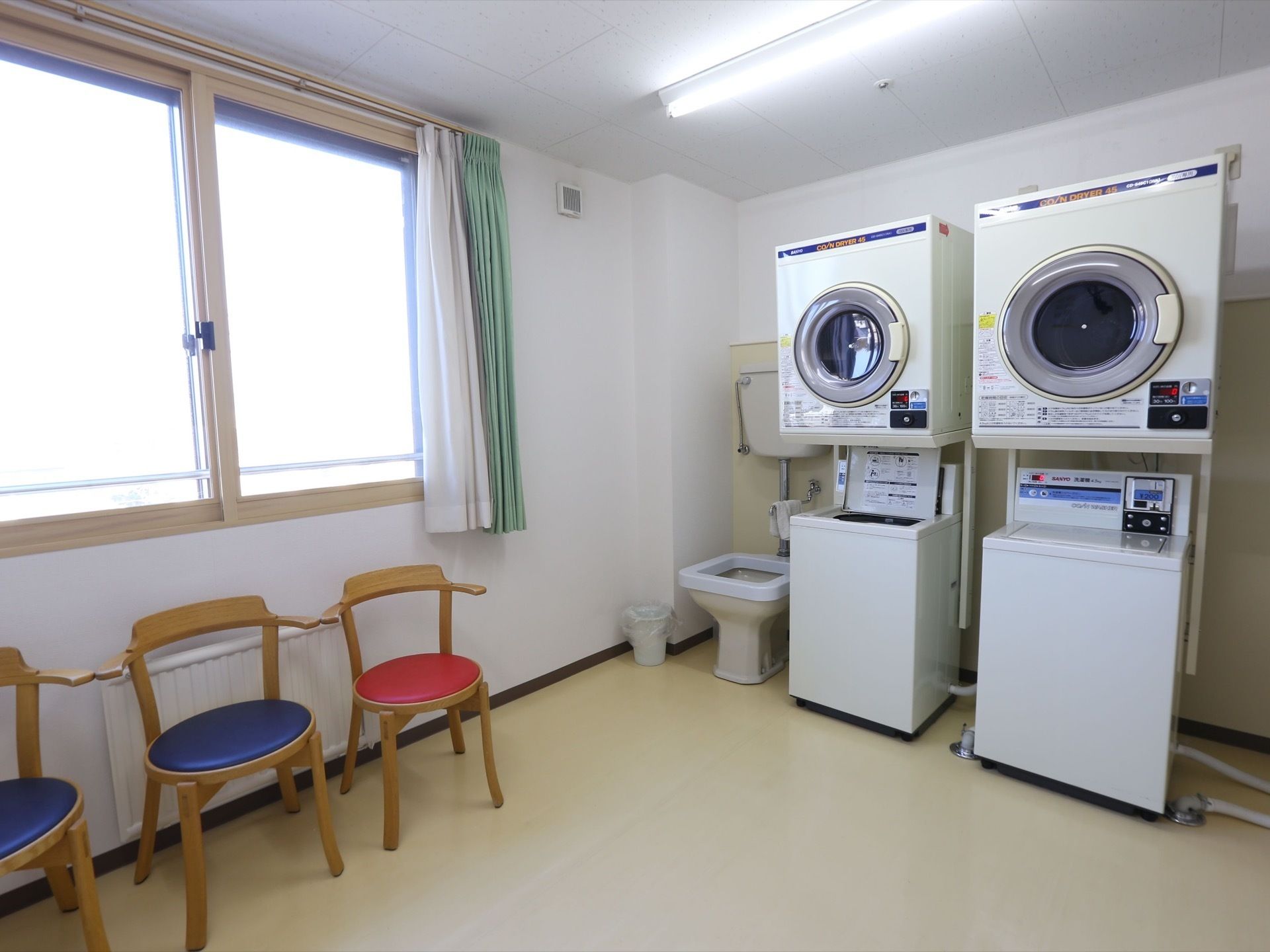 明るいグレーの天井に白の壁、ベージュの床である。乾燥機と洗濯機が２台ずつ設置され、座面が赤と青の椅子が置かれている。