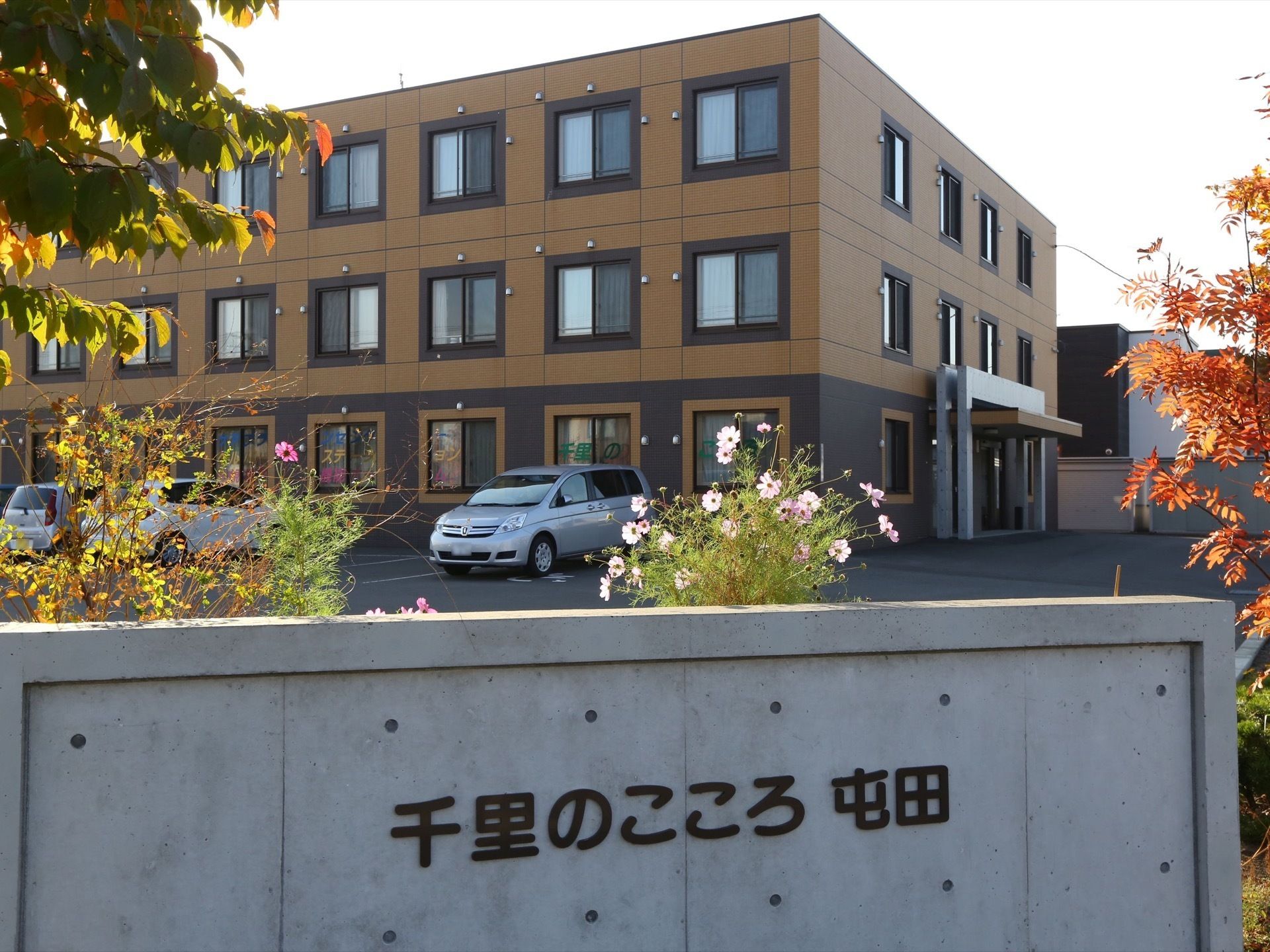 外壁や窓枠が茶系で統一された、落ち着いた外観の施設である。看板の周りには、色とりどりの植物が植えられている。