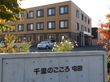 外壁や窓枠が茶系で統一された、落ち着いた外観の施設である。看板の周りには、色とりどりの植物が植えられている。