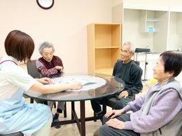 共有スペースで丸テーブルを囲みおしゃべりを楽しむ3人の入居者とエプロン姿のスタッフ。