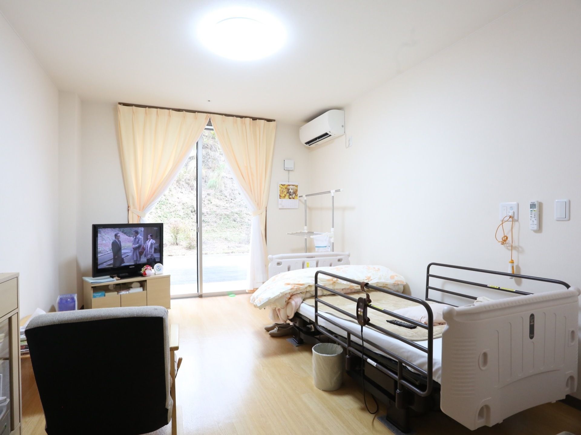 シンプルな作りの居室に、介護用ベッドやテレビ、椅子などが置かれている。エアコンや呼び出しボタンが備えつけられている。