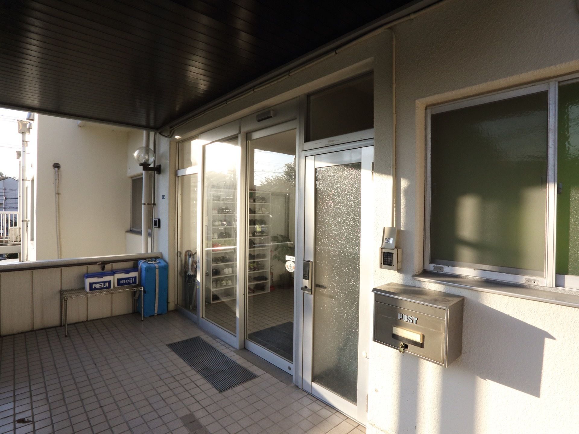 ガラス張りの自動ドアのある施設の玄関。床はタイルになっている。壁には窓がついており、郵便ポストがある。
