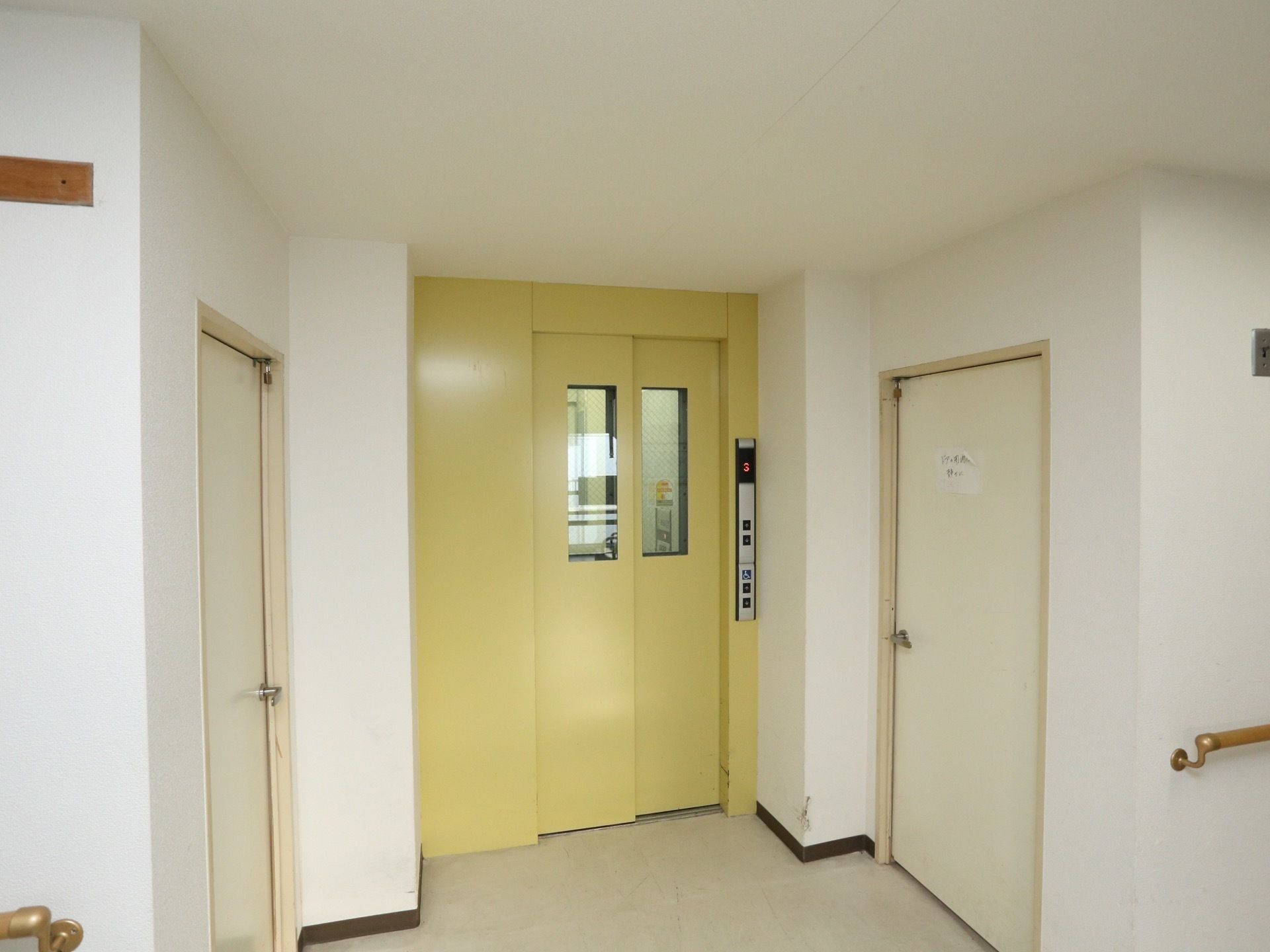 クリーム色の扉のエレベーターがある。ガラスののぞき窓がついている。すぐ横には非常階段へと続く扉がある。