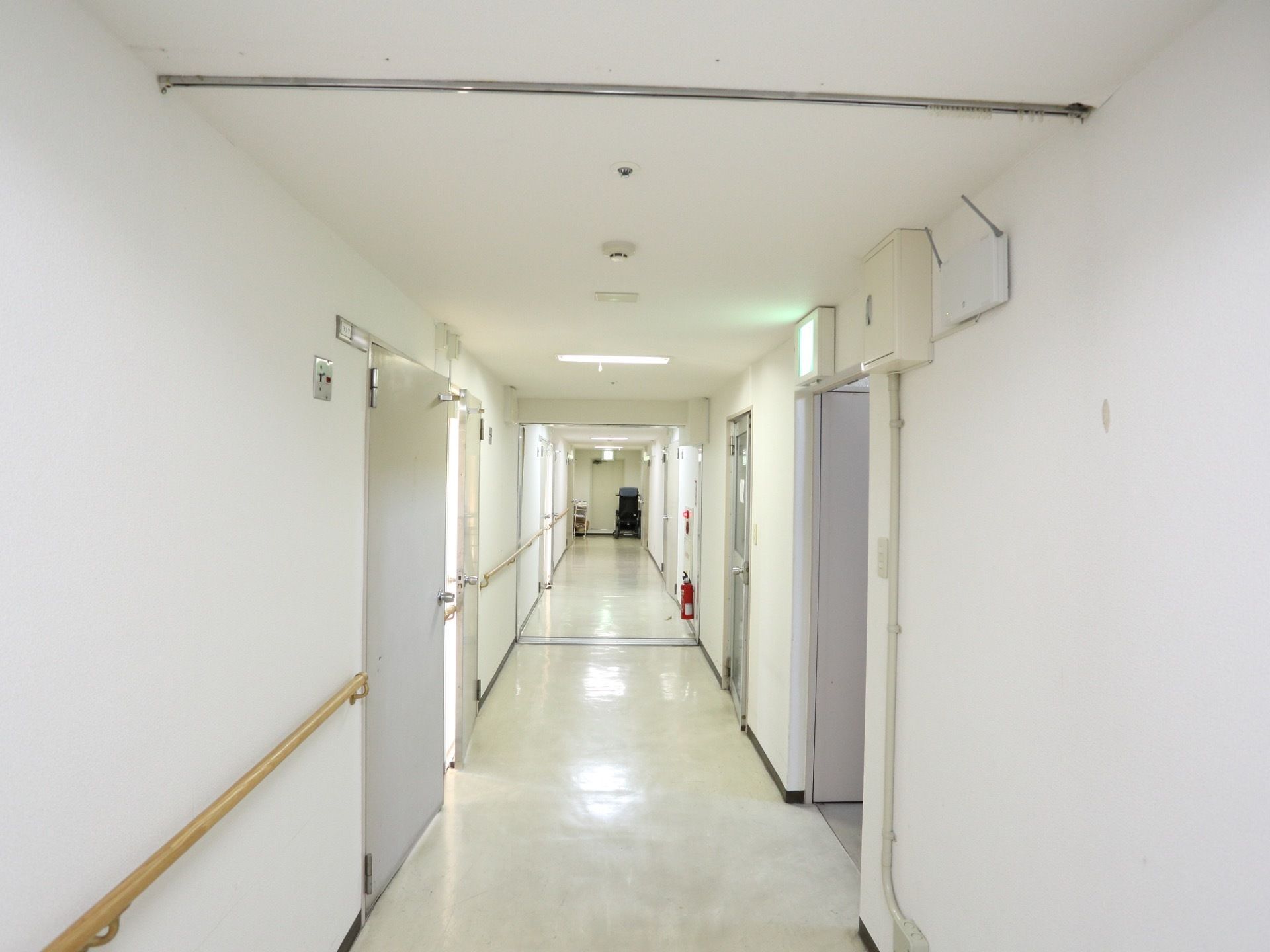白い壁と入居者様の居室のドアが並ぶ長い廊下。照明は明るく、清潔感がある。壁には手すりが設置されている。