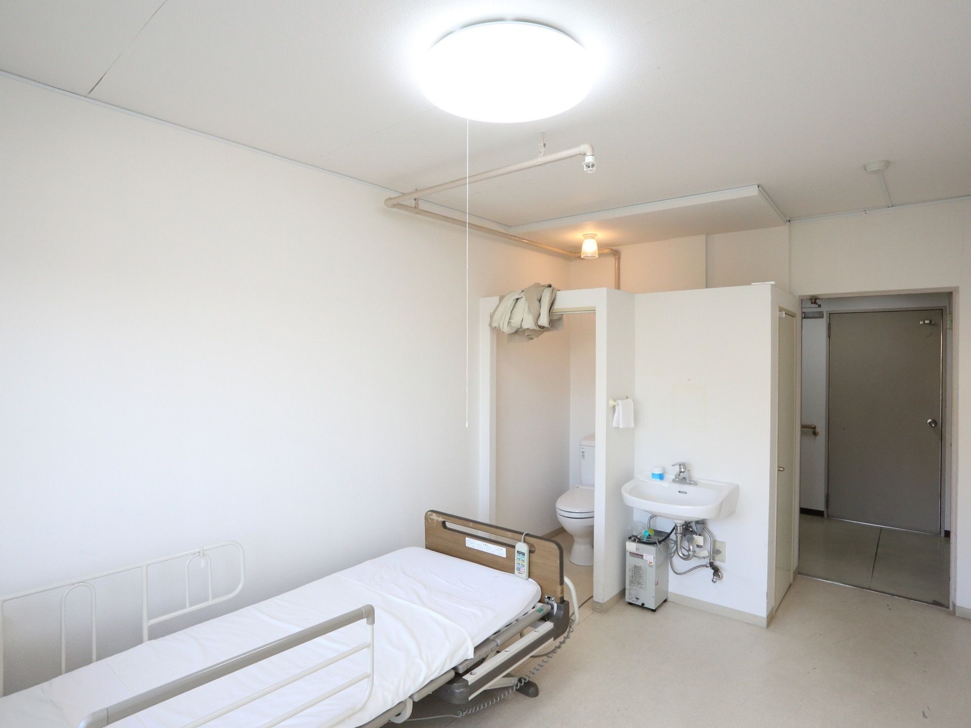 白い壁と明るい照明の入居者様の居室。洗面台とトイレがある。柵のついた介護用ベッドが置かれている。天井が高く、広々としている。