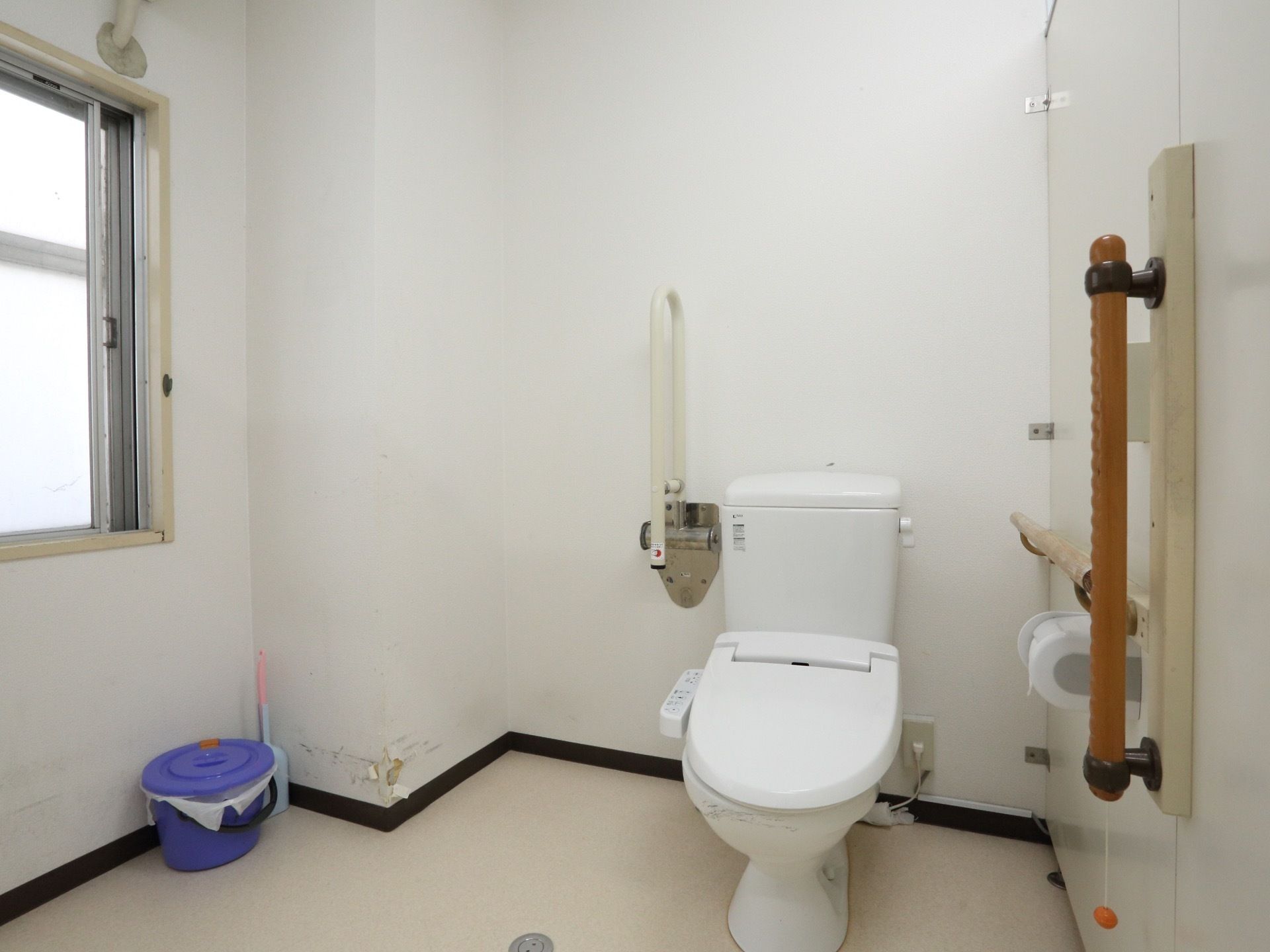 大きな窓がついている、広い個室のトイレ。壁には縦横方面に手すりがついており、便座には可動式のひじ掛けが設置されている。