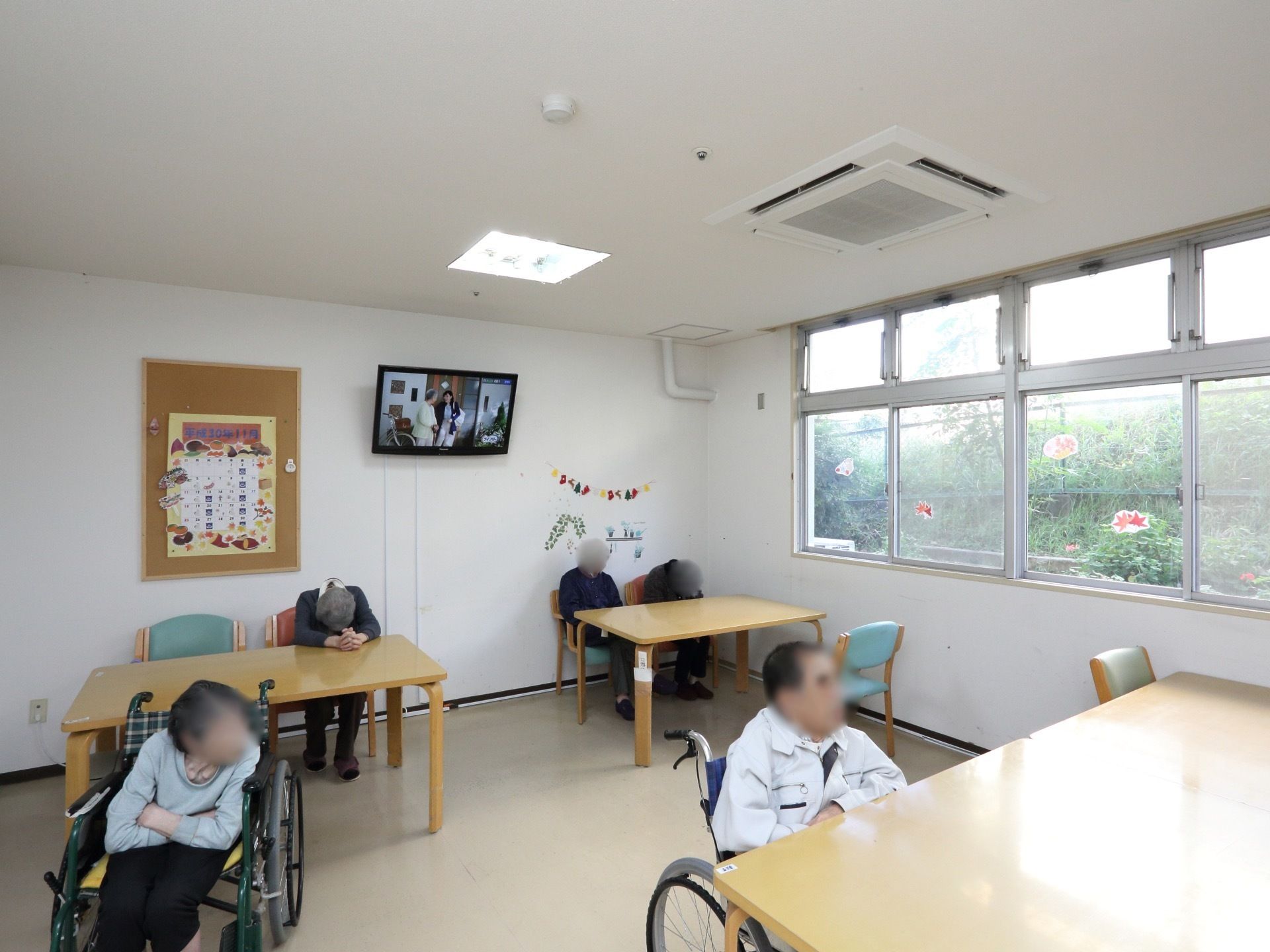 大きな窓から緑の植物と花が望める食堂がある。テーブルと椅子が並び、壁の高い位置には薄型テレビが設置されている。