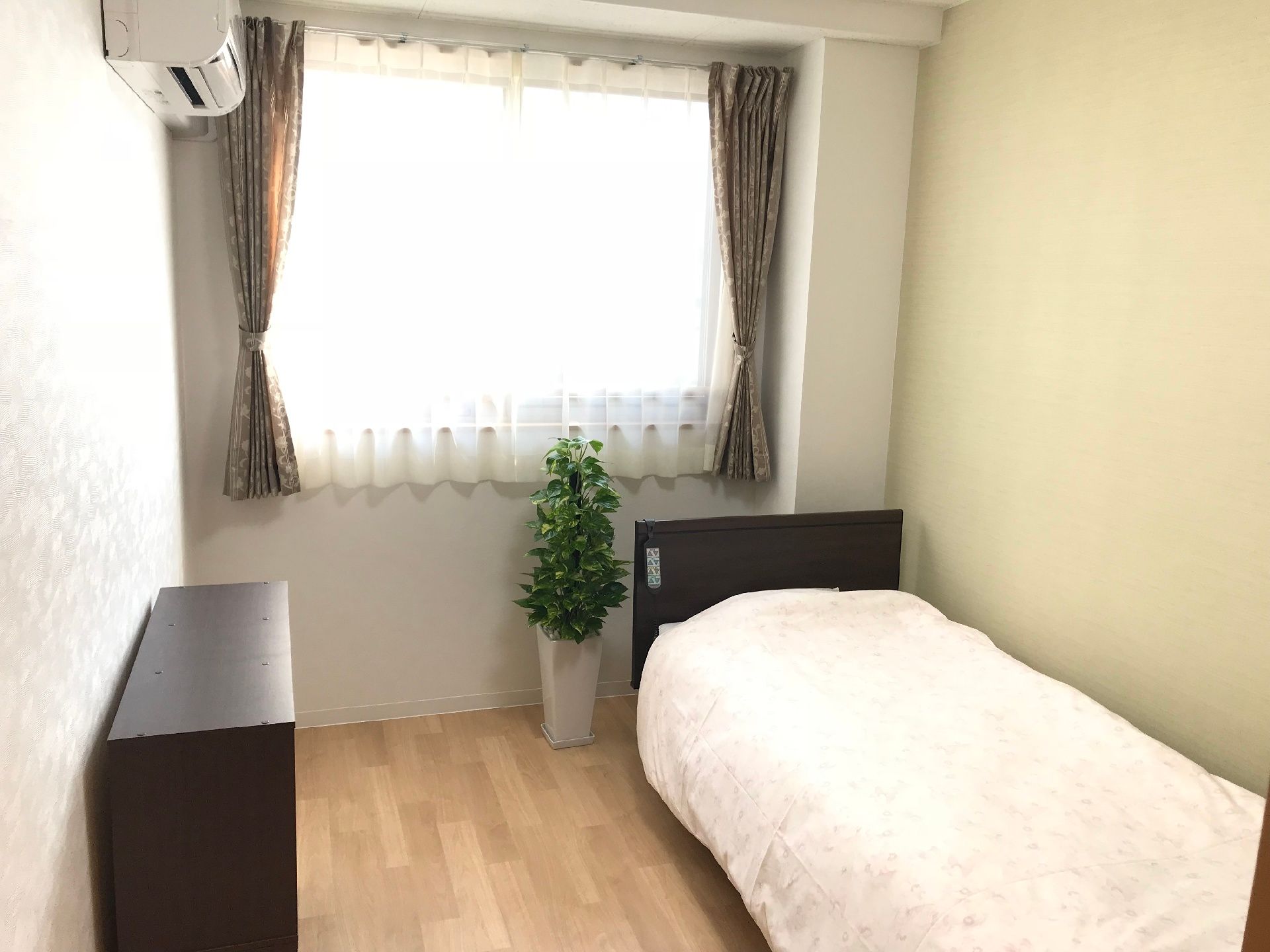 窓から差し込む光が印象的な部屋。ベッドとエアコン、タンス、観葉植物が見える