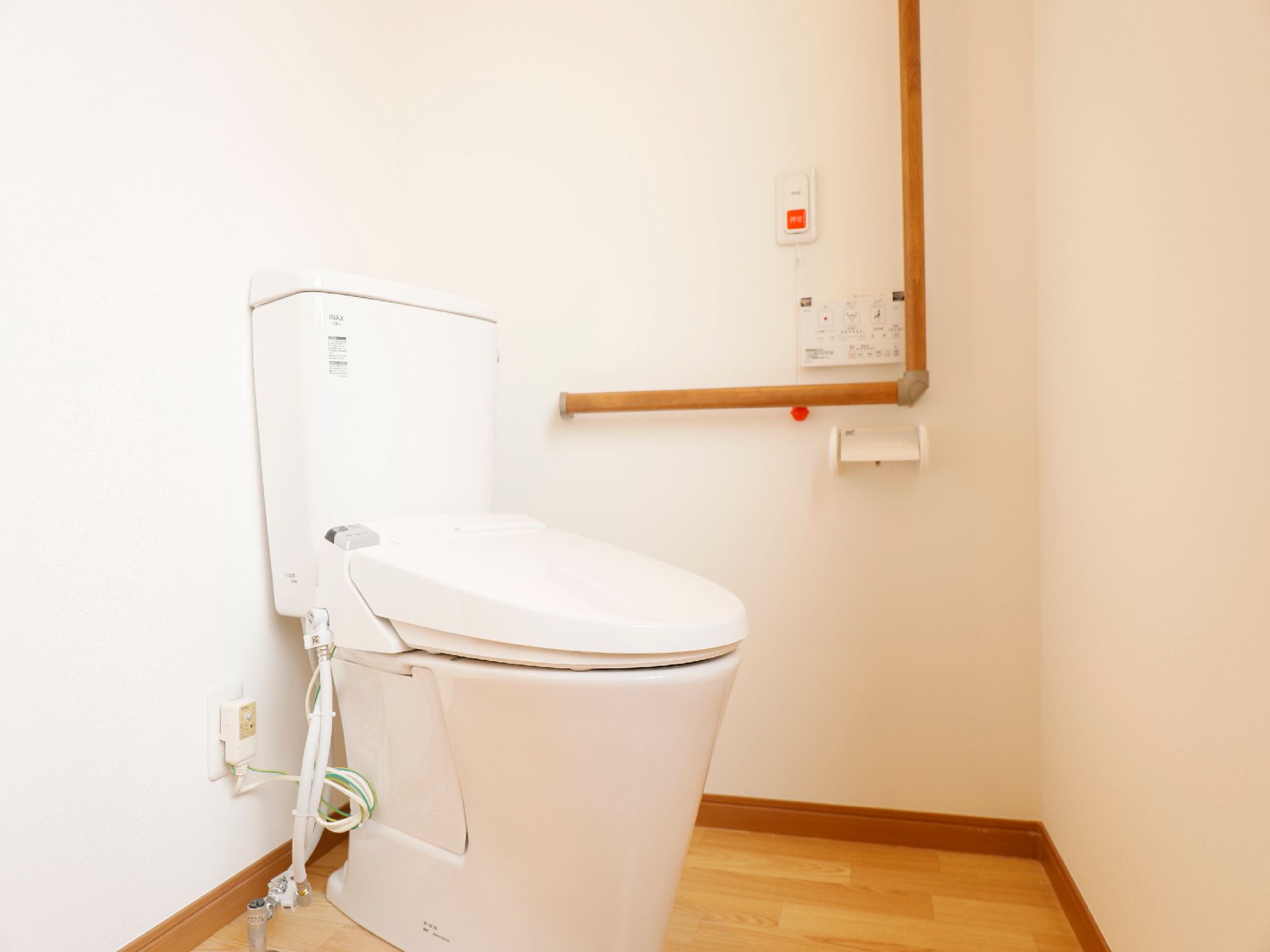 タンク付きの白い洋式トイレがある。壁に紐と玉がついたナースコールと逆Ｌ字型の手すりが設置されている。