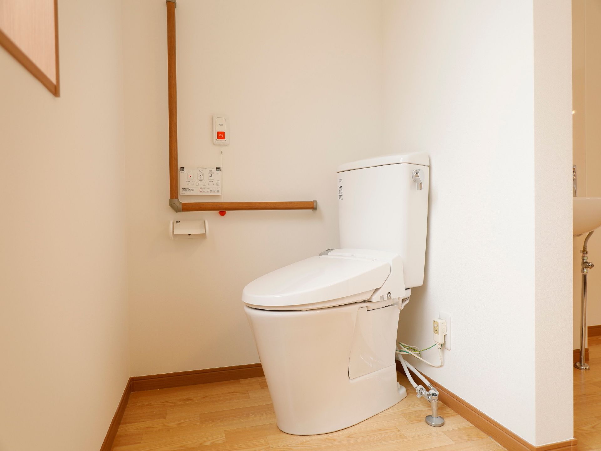 白いタンクに洗浄レバーがついた洋式トイレがある。壁に手すりと温水洗浄便座のリモコン、ペーパーホルダーが設置されている。