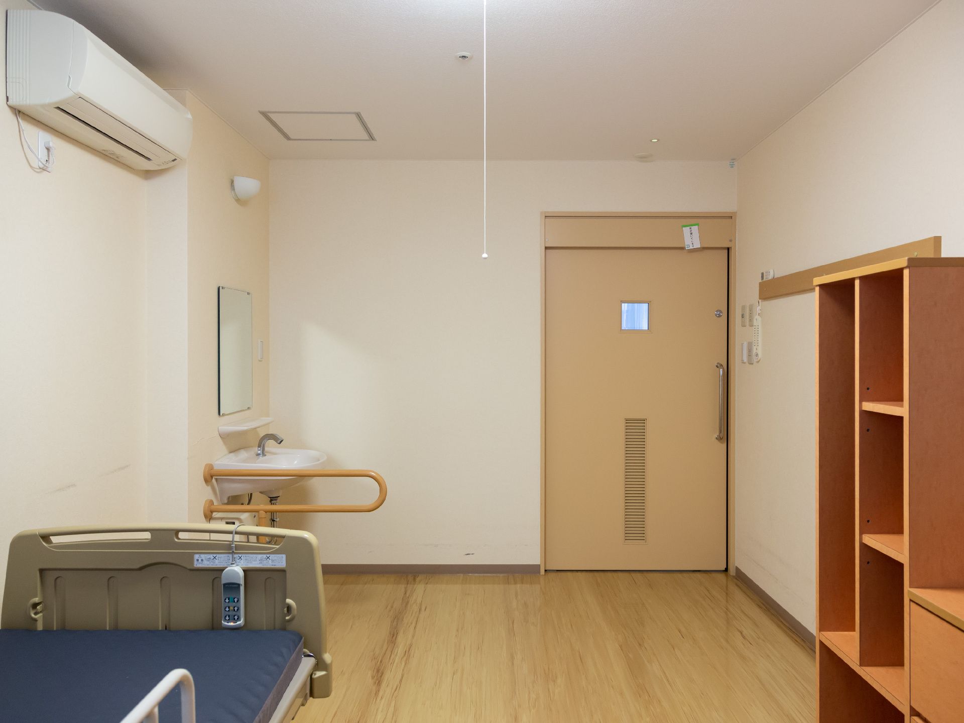 居室には介護用ベッドが壁際に置かれ、壁にベッドライトが取りつけられている。天井近くにエアコンが設置されている。