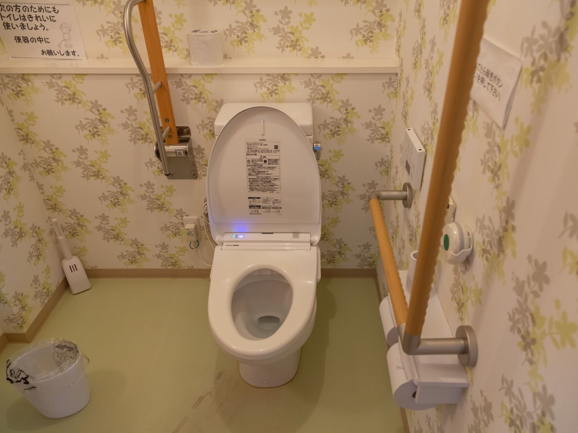 温水洗浄便座の洋式トイレがある。壁紙には植物が描かれている。便座左側の壁にＬ字の手すりと緊急ボタンがある。