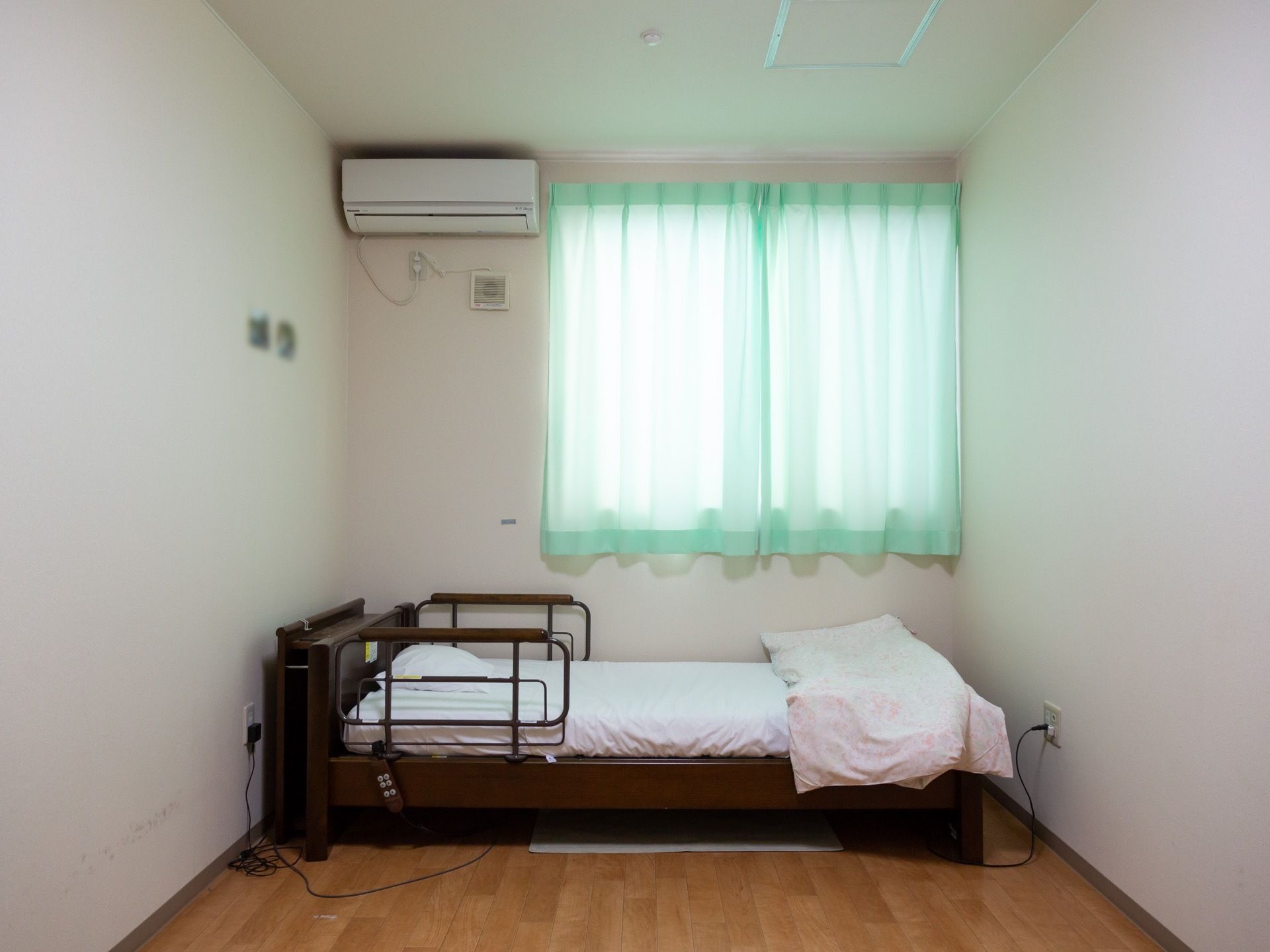 木目調のフローリングに、白い壁の居室である。壁にはエアコン、床には介護ベッド、窓にはグリーンのカーテンが掛かっている。