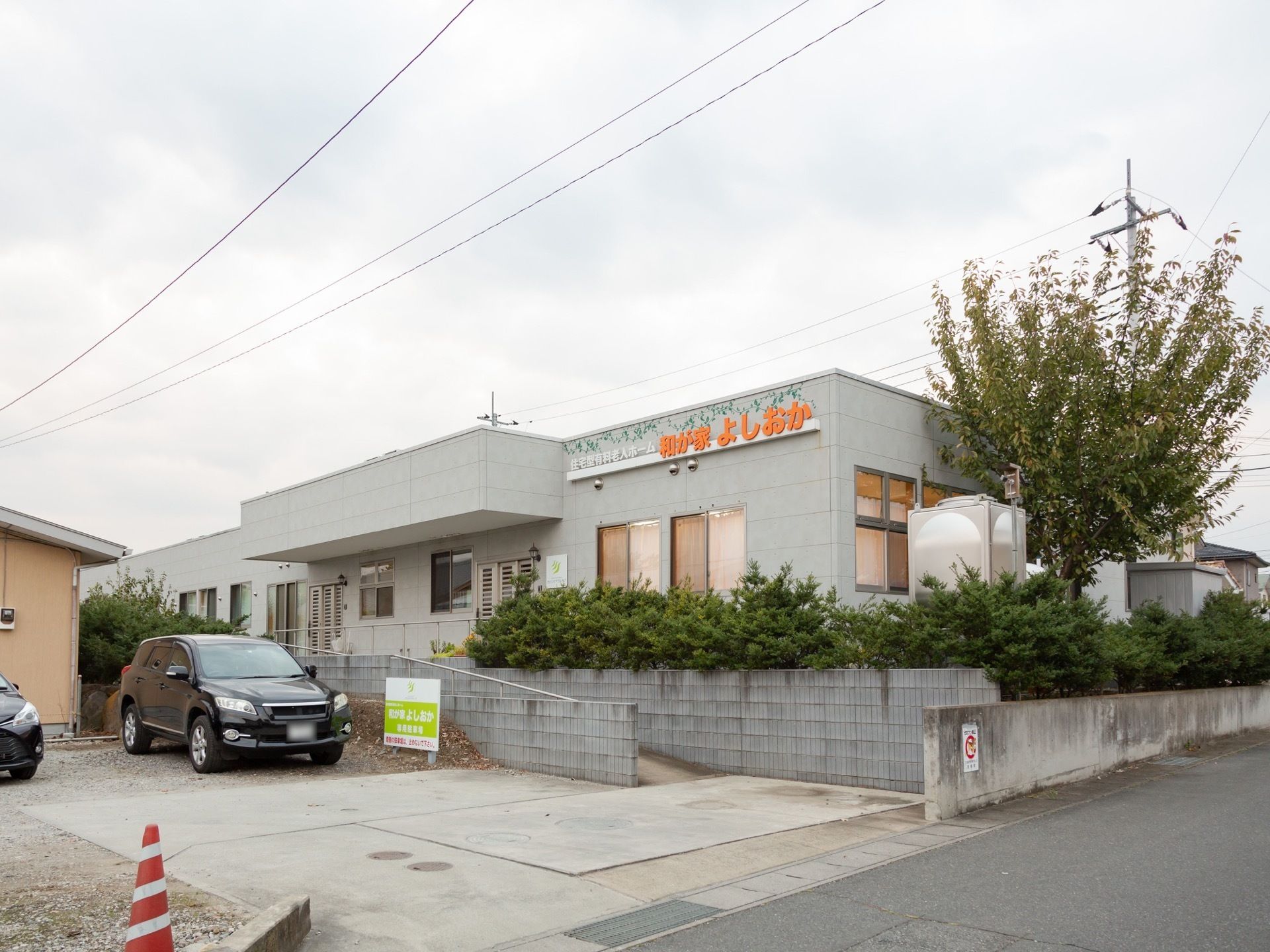 １階建てグレーの施設建物である。駐車場から玄関まではスロープになっていて、建物の周りには植栽が植わっている。