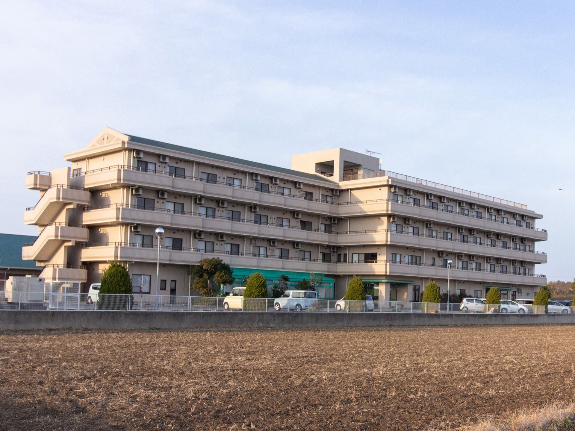 ベージュを基調とした落ち着いた色合いの建物。４階建てとなっており、非常階段が設置されている。手前には畑が広がっている。