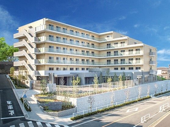 日当たりのいい場所に建っているベージュ系の外壁の大きな建物。敷地内には駐車場があり、草木が植えられている。