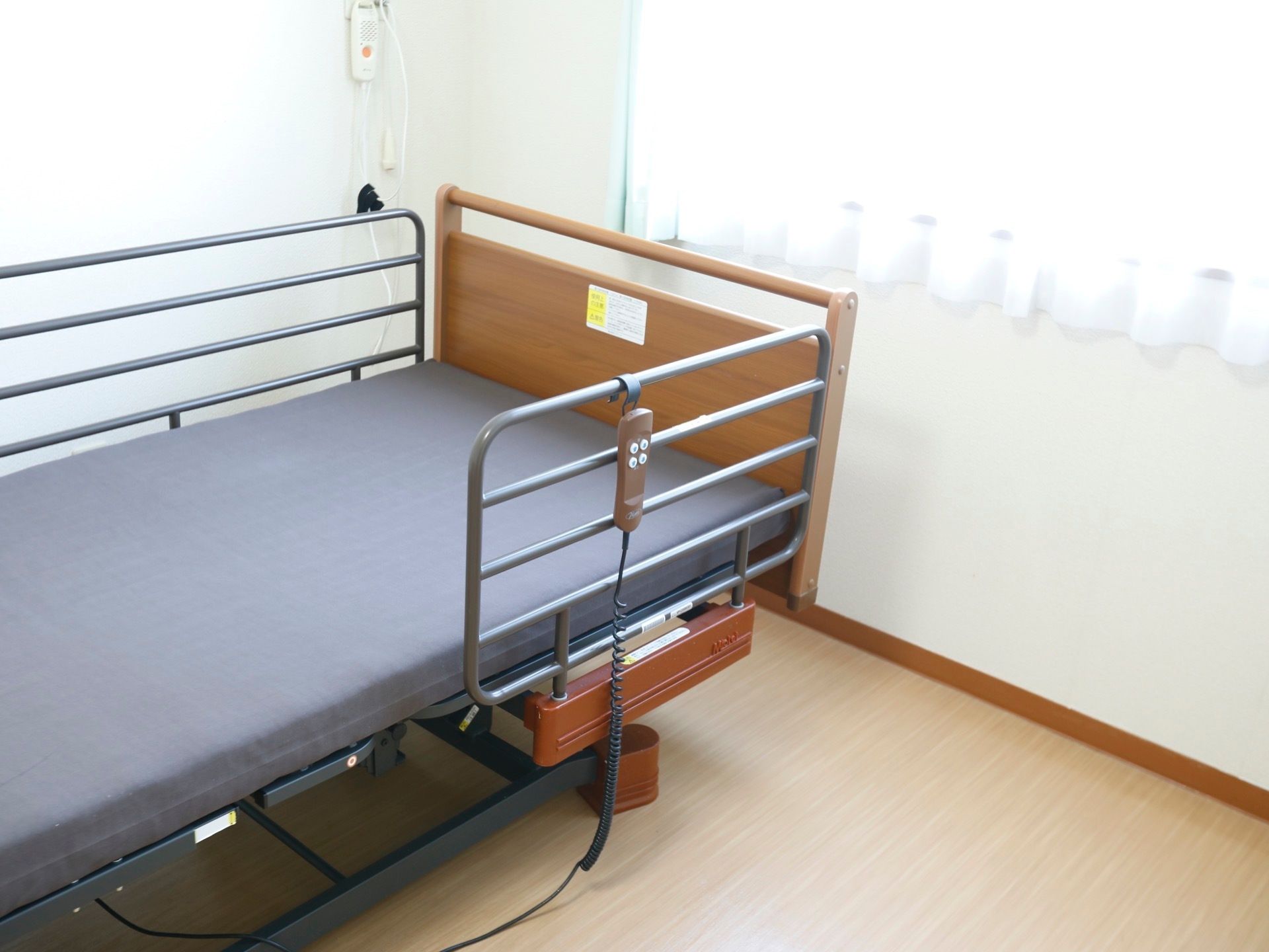 居室の窓際に、介護用ベッドが配置されている。腰高窓があり、日差しが差し込んでいる。壁にはナースコールを完備。