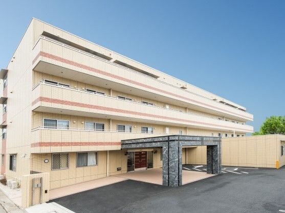 ベージュ系の外壁の4階建ての建物の敷地内には駐車場もあり、各フロアにベランダがある。