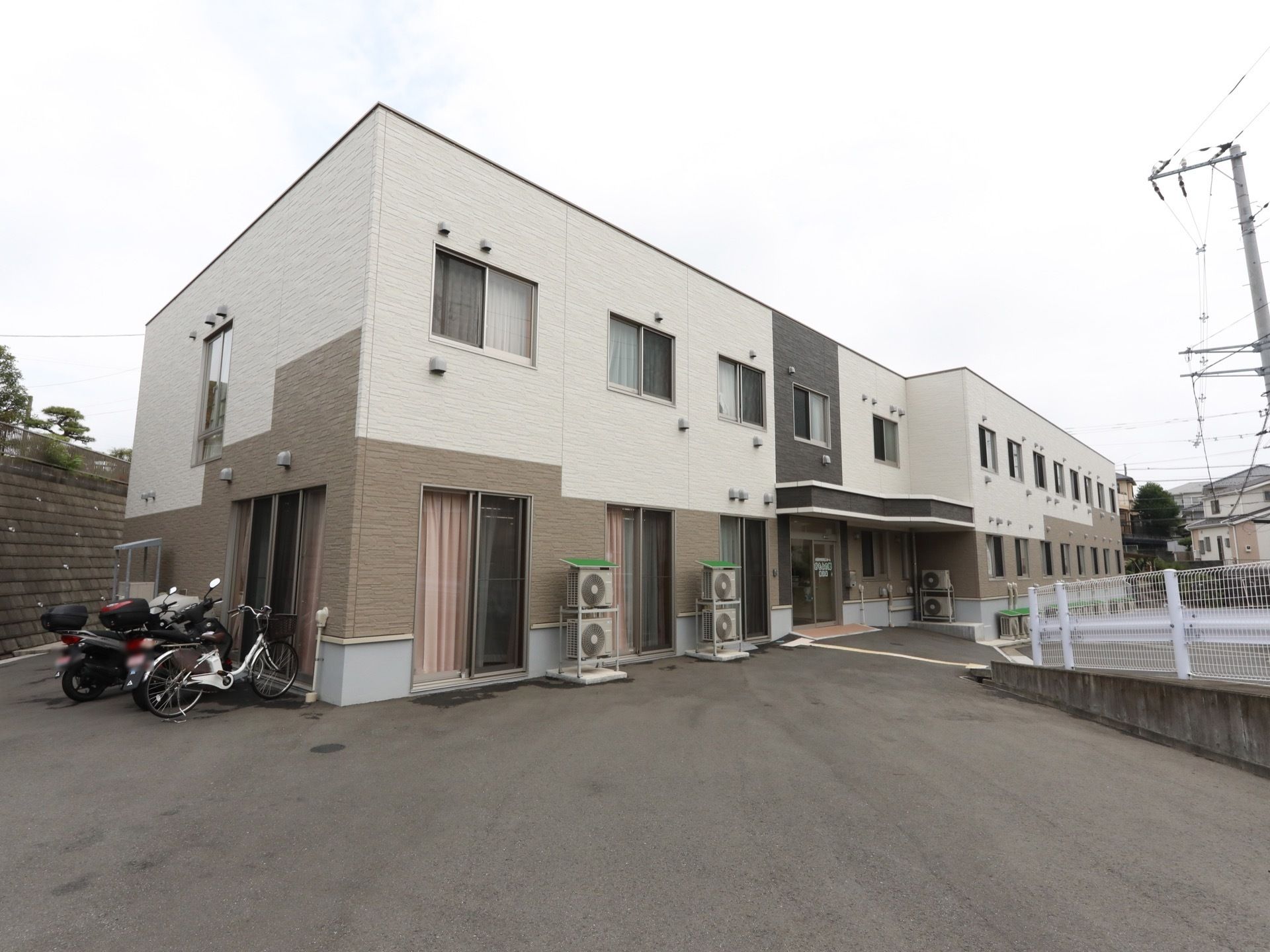 横幅の広い2階建ての建物が、住宅街の一角に建っている。建物の左側が舗装され、オートバイや自転車が停められている。