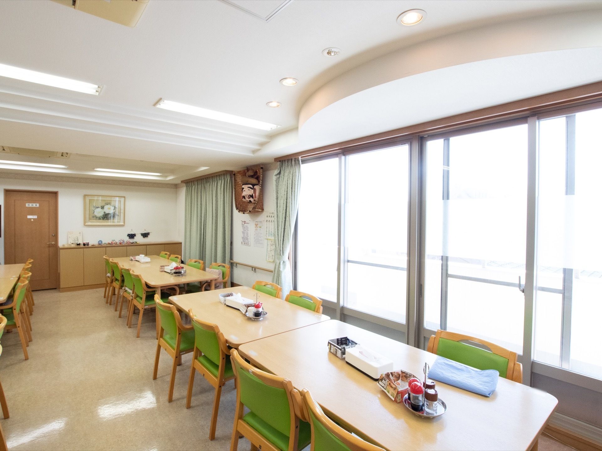 たくさんのテーブルと椅子が並ぶ食堂。壁には大きな窓が複数あり、日光が室内に差し込んでいる。テーブルの上には調味料入れなどが置かれている。