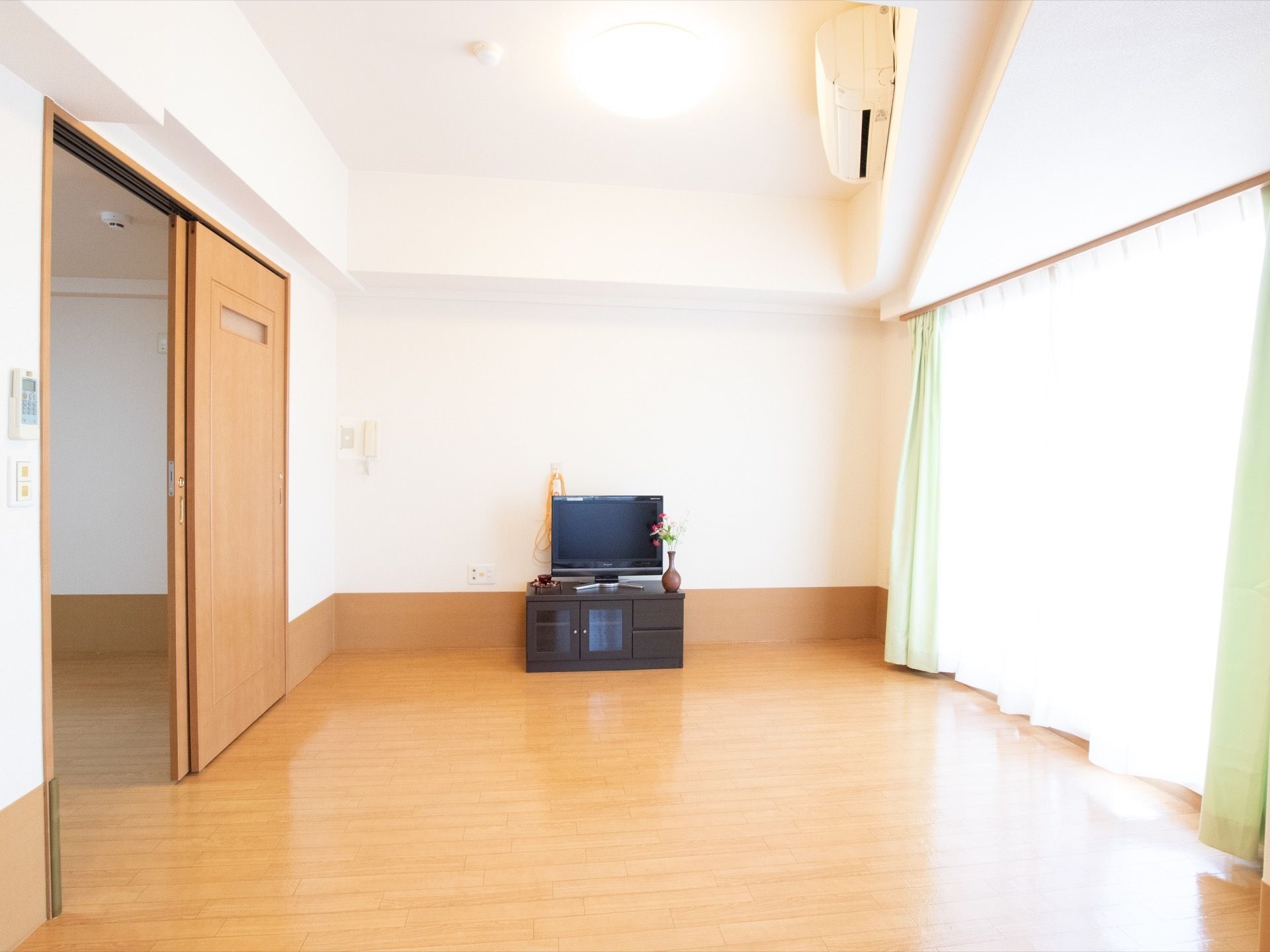 広いフローリングの床の居室に、テレビとテレビ台が置かれている。右側には大きな窓があり、左側には他の部屋へと続く入口がある。