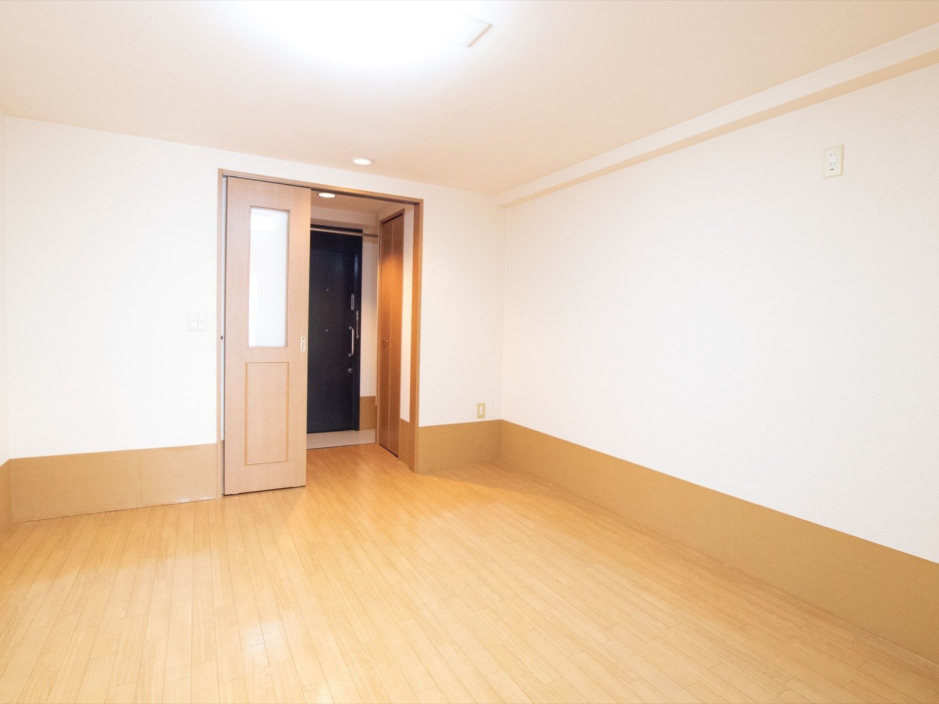 広い洋室があり、正面の奥には部屋の入口である引き戸の扉が設けられている。その更に奥には、玄関のドアが見える。
