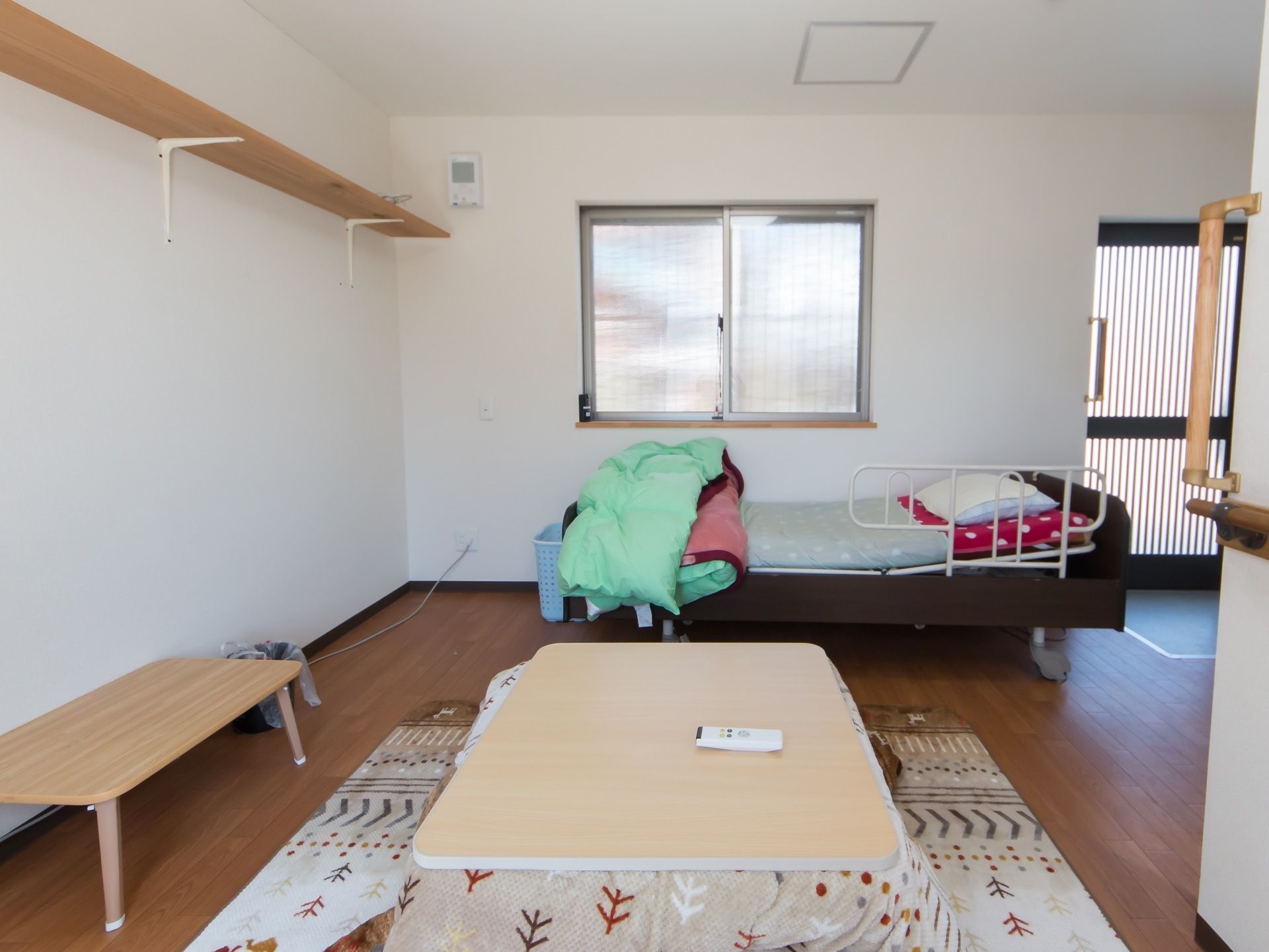 広々としたフローリングの居室である。部屋の中央にはこたつが置かれており、奥にある腰高窓の側には介護ベッドが設置されている。