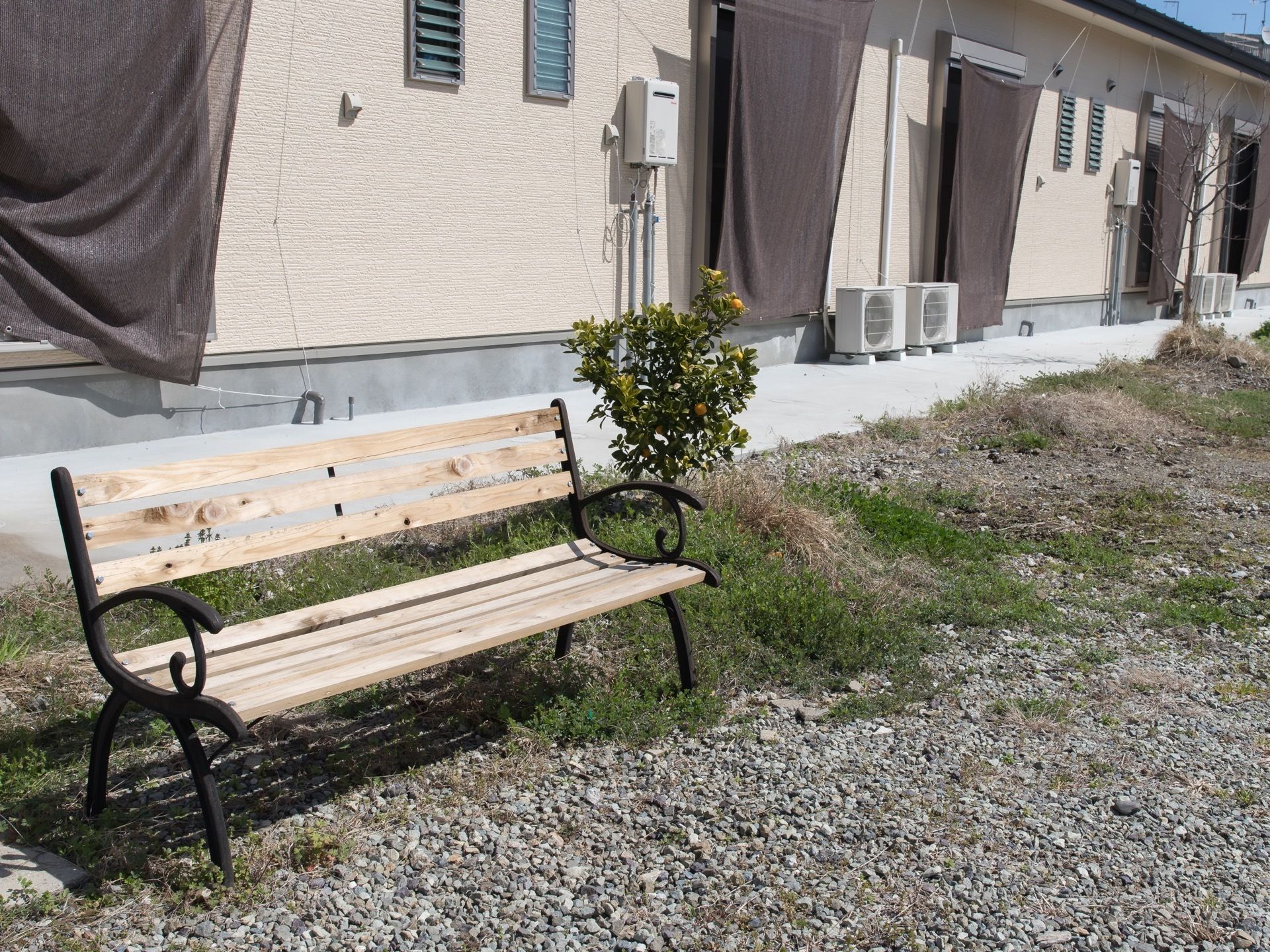 庭には木製のベンチが置かれており、横には小さな木が植えられている。後ろにある建物の窓には日よけが付けられている。
