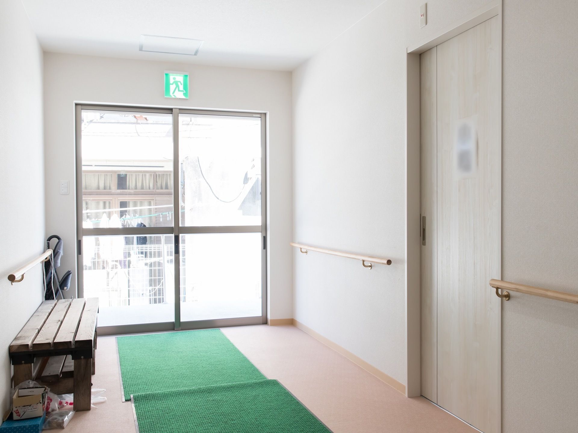 エントランスの壁には手すりが設置されており、左側にはベンチが置かれている。入口はガラスの引き戸になっていて、光が入る明るい空間である。