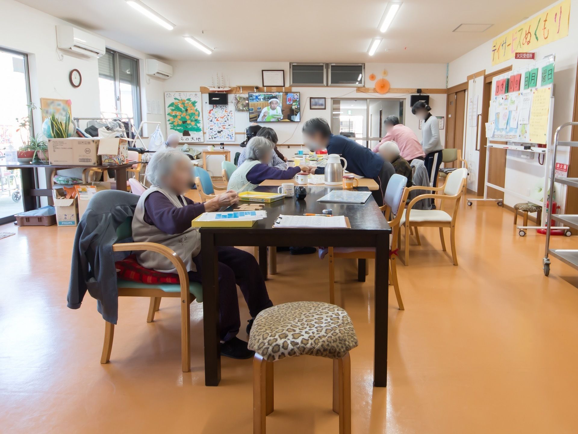 共有スペースにはテーブルが設置されており、入居者が座って話などをしている。奥の壁にはテレビが掛けられている。