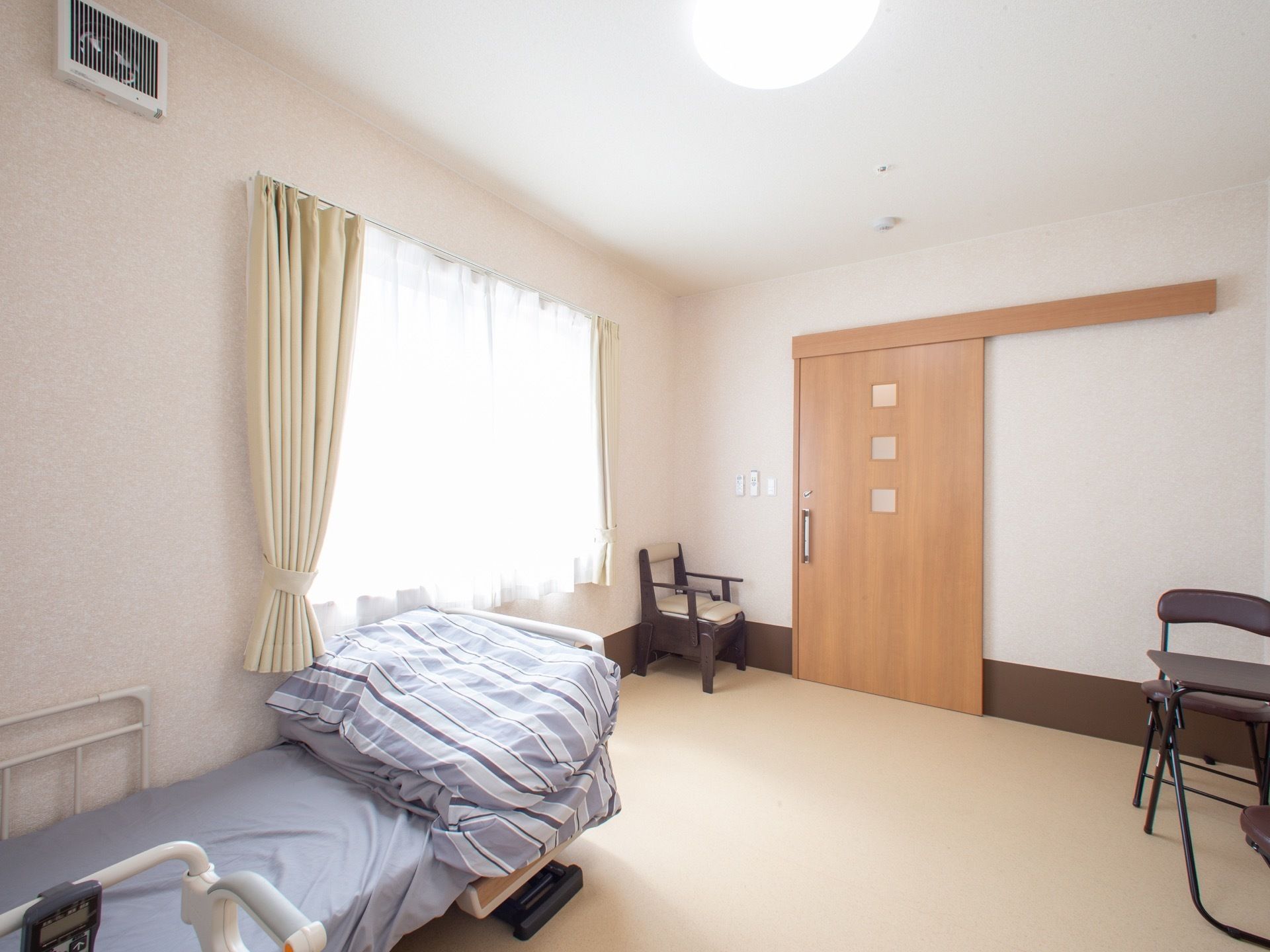明色系に統一された居室である。扉は引き戸式タイプ。大きな窓も備え付けられている。ベッドも置かれている。