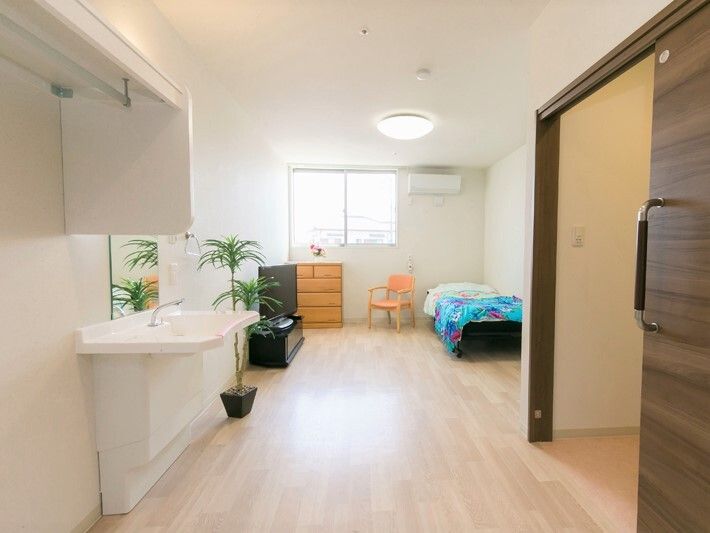 右側にはトイレのスライドドアがあり、左側には車いす対応洗面台が付き、フローリングの居室にはベッドやタンス、観葉植物などが置かれている。