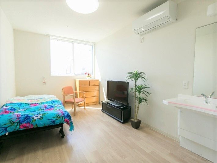 エアコンが付いた白を基調とした部屋には車いす対応洗面台やベッド、タンス、いす、テレビ、観葉植物などが置かれ明るい雰囲気。