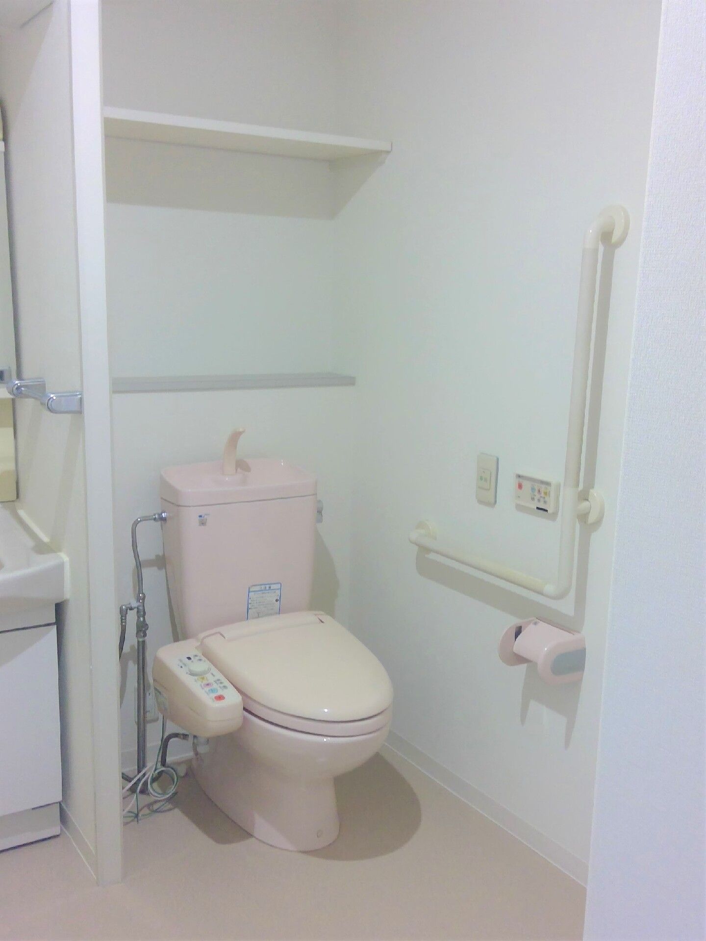エル字型の手すりや呼び出しボタン、トイレットペーパーホルダーが設置された壁。トイレは温水便座で便座に操作ボタンが付いている。