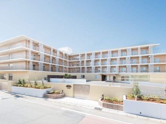 エル字型で各フロアにベランダがあるベージュの外壁の建物。門のそばには草木が植えられている。
