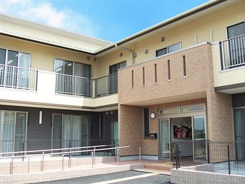 エル字型の建物は２階建てで玄関には手すり付きのスロープがあり、青い空が広がっている。
