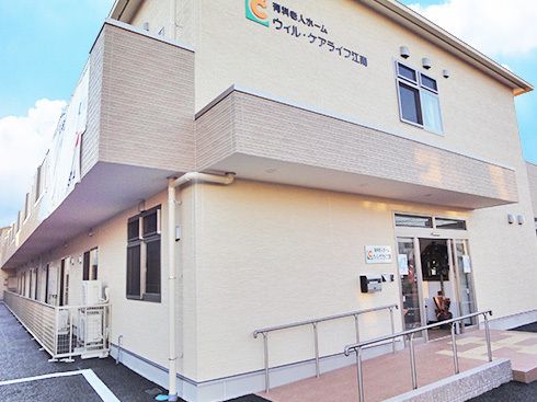 ベージュ系の2階建ての建物。玄関には屋根が付き、手すり付きのスロープもある。