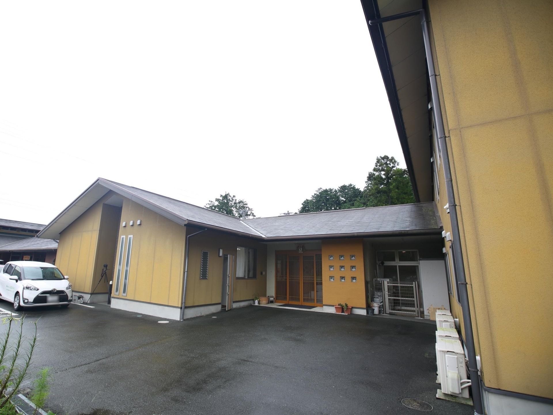 落ち着いたベージュ調に施された施設外観である。車が1台停められており、玄関にはマットが敷かれている。