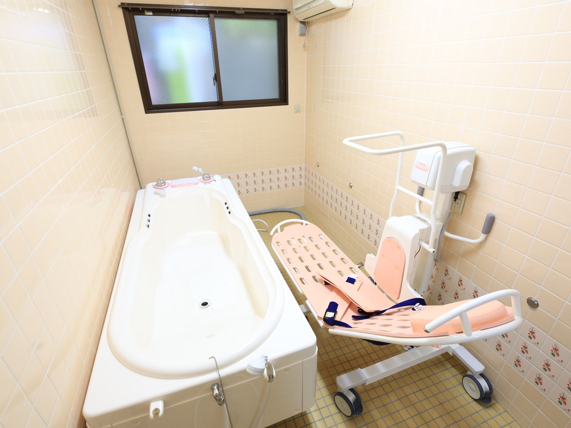可動式のストレッチャーの横に白色の大きな浴槽がある。窓近くの壁の上部にエアコンがある。すりガラスの窓がある。