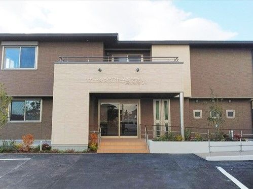 ブラウン系の建物は2階建てで駐車場があり、玄関には手すり付きのスロープが付いていて、2階部分にはテラスもある。