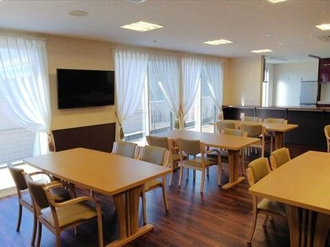 大きな壁掛けテレビが設置された食堂には4人掛けのダイニングテーブルが並び、レースカーテンとカーテンが窓に付いている。