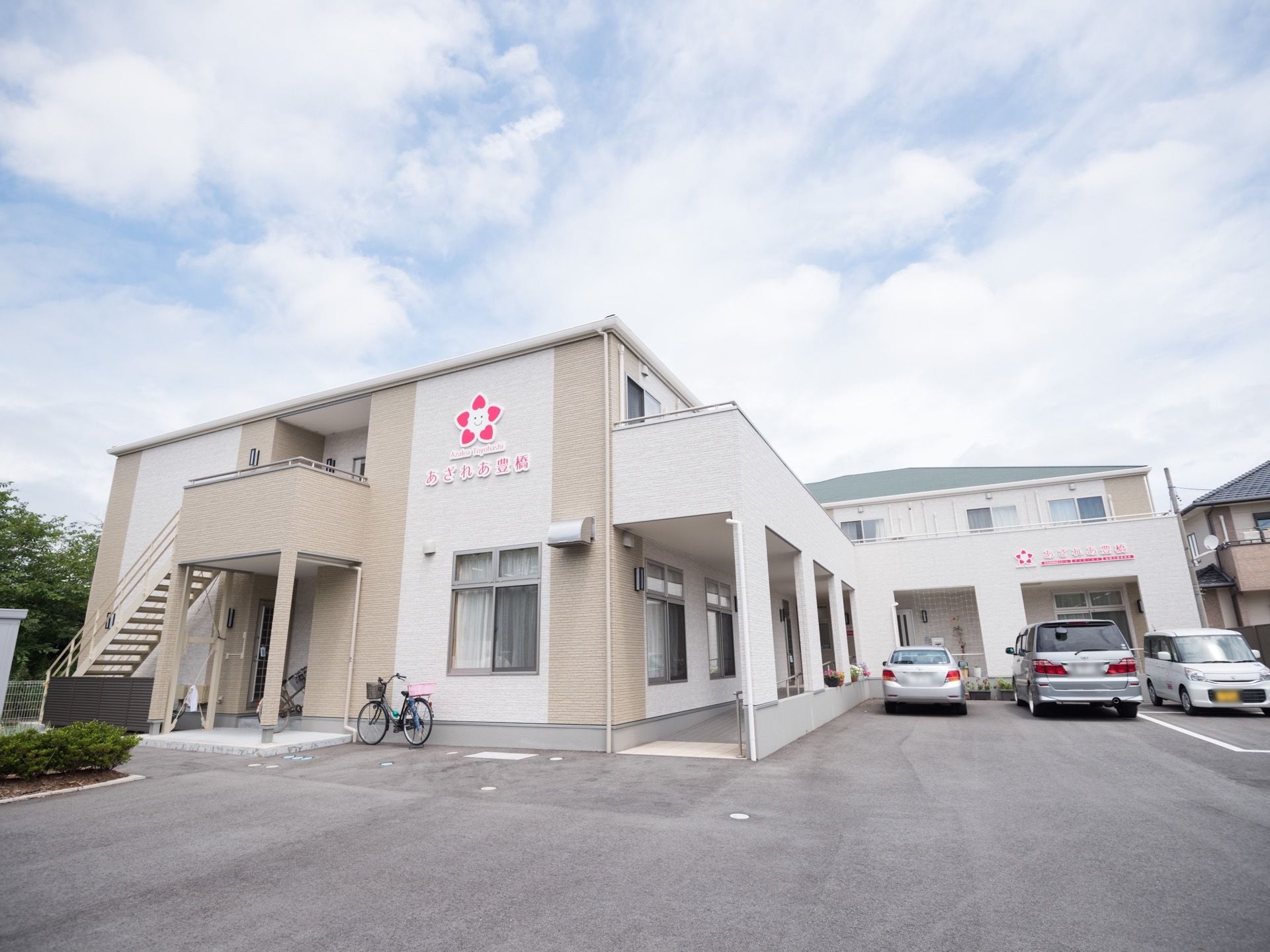 ２階建てのＬ字型の施設の周囲に、アスファルトで舗装されたスペースが広がっており、複数の自動車が駐車されている。