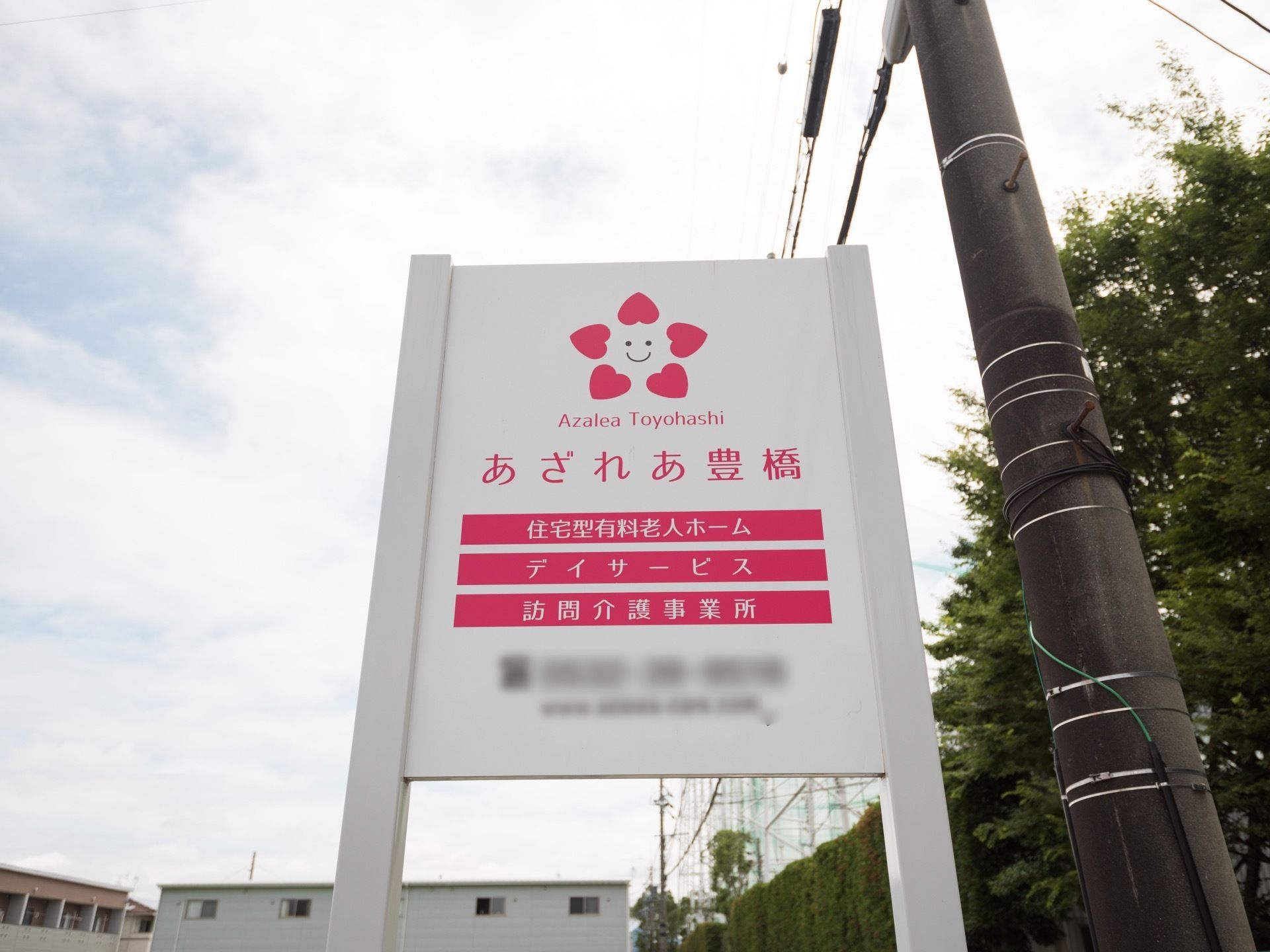 電柱の左側に白い看板が設置されている。施設名称やシンボルマーク、主なサービス内容がわかりやすく表示してある。