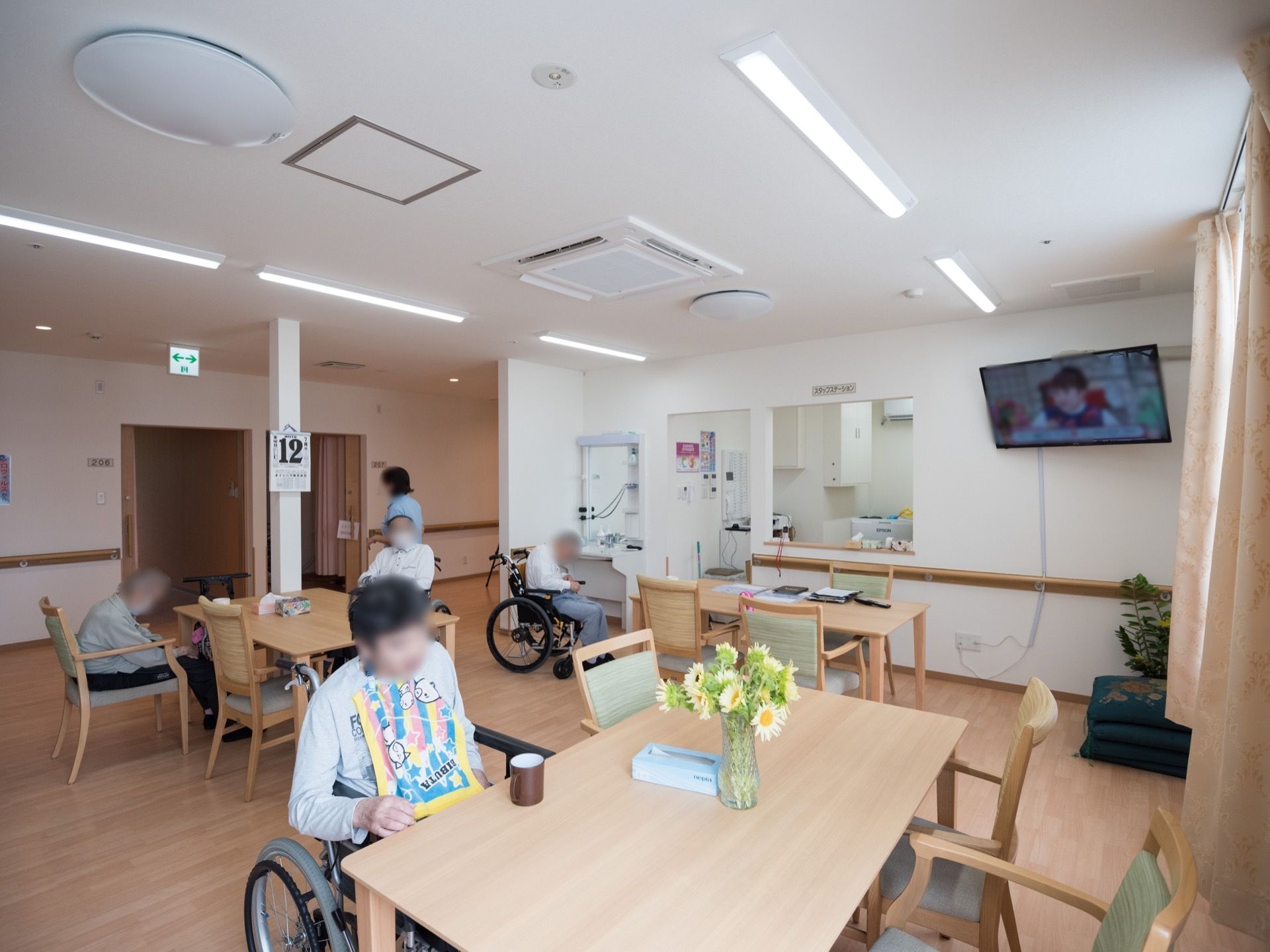 明るく清潔感のある食堂で過ごす人々の姿が見える。切り花が生けられた花瓶や観葉植物の鉢植えが用意されている。